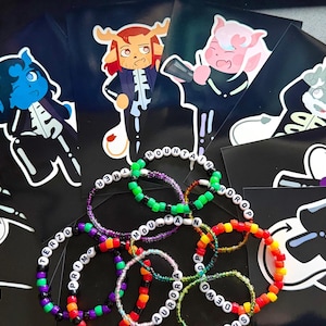 May include: A collection of colorful beaded bracelets with white letter beads spelling out words like "MOUNTAIN" and "AURORA" are displayed with several character stickers on black backgrounds. The stickers feature cartoon characters with skeleton designs.