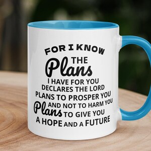 Puede incluir: Taza de cerámica blanca con interior y asa azules. La taza presenta el texto "FOR I KNOW THE Plans" y un versículo bíblico. La taza está sobre una superficie de madera.