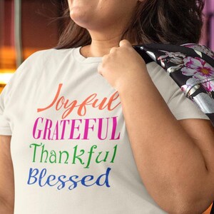 May include: A cream-coloured short-sleeve t-shirt featuring the text Joyful in orange, GRATEFUL in magenta, Thankful in green, and Blessed in blue. The shirt is paired with black leggings and a black jacket with a pink and white floral pattern.