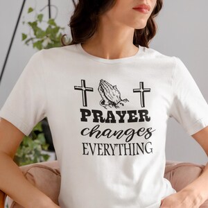 Puede incluir: Una camiseta blanca de manga corta con un estampado gráfico en negro. El diseño incluye dos cruces a los lados de unas manos en oración, con el texto PRAYER changes EVERYTHING impreso en una combinación de letras negritas y cursivas.