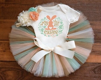 May include: A white baby onesie with the text "My 1st Easter" and a bunny silhouette. The onesie is paired with a multi-colored tulle tutu and a floral headband.