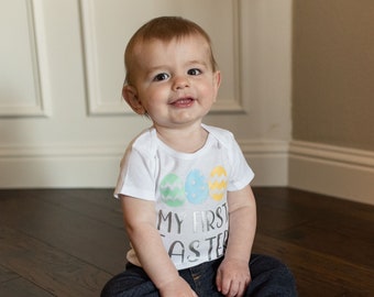 Puede incluir: Un bebé con un body blanco con el texto "My First Easter" y tres huevos de colores. El bebé está sentado en un suelo de madera y sonriendo a la cámara.