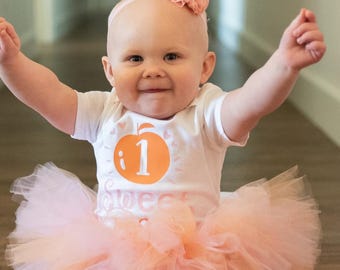 Peut inclure: Un body blanc à manches courtes orné d'un motif orange représentant une pêche avec le chiffre un à l'intérieur et le mot sweet en dessous, associé à un tutu en tulle pêche clair et un bandeau accessoire avec une petite couronne rose.