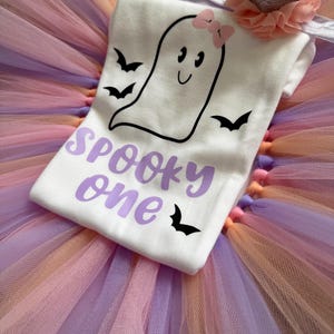 May include: A white short-sleeved shirt featuring a black ghost graphic with a pink bow, black bats, and the purple text "SPOOKY one" layered over a multi-coloured tulle tutu skirt with a silver glitter crown and a peach-coloured fabric flower.