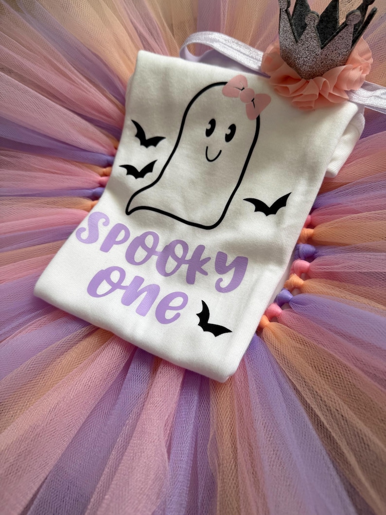 May include: A white short-sleeved shirt featuring a black ghost graphic with a pink bow, black bats, and the purple text "SPOOKY one" layered over a multi-coloured tulle tutu skirt with a silver glitter crown and a peach-coloured fabric flower.