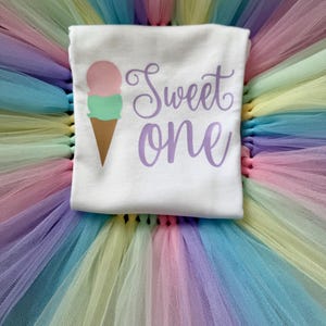 May include: A white t-shirt featuring a graphic of a two-scoop ice cream cone with a pink top scoop and a mint green bottom scoop. The text "Sweet one" is printed in a purple cursive font to the right of the ice cream cone graphic.