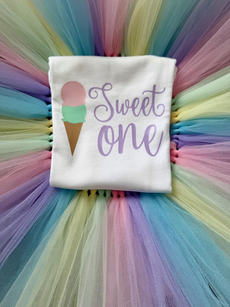 May include: A white t-shirt featuring a graphic of a two-scoop ice cream cone with a pink top scoop and a mint green bottom scoop. The text "Sweet one" is printed in a purple cursive font to the right of the ice cream cone graphic.