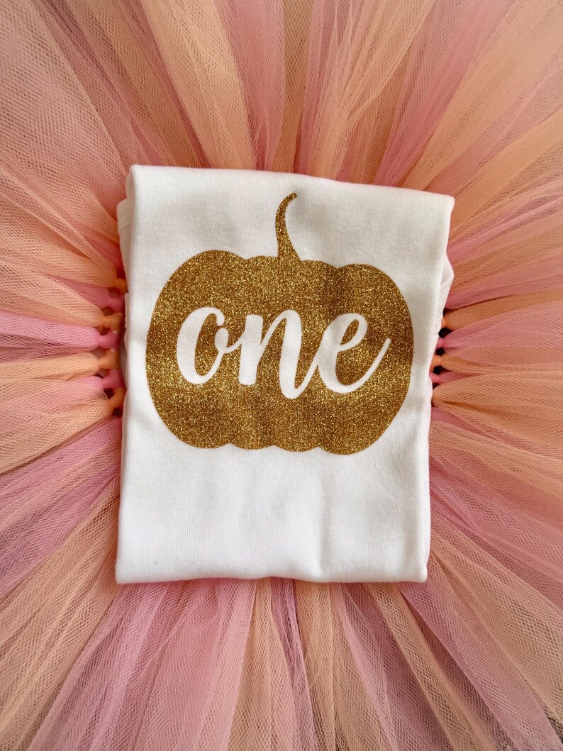May include: A white t-shirt featuring a gold glitter pumpkin graphic with the word one written in white script across the center, placed on top of a layered peach and light pink tulle skirt with a gathered texture and sheer fabric finish.
