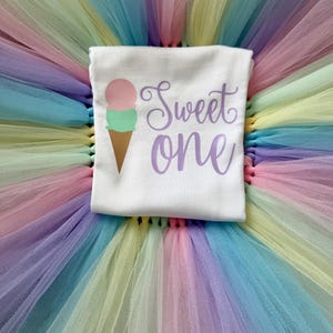 May include: A white short-sleeved shirt featuring a graphic of a two-scoop ice cream cone in pink and green with the text Sweet one in purple script, placed on a layered tutu skirt made of pastel-colored tulle in shades of pink, yellow, blue, and green.