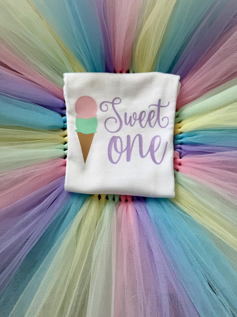 May include: A white short-sleeved shirt featuring a graphic of a two-scoop ice cream cone in pink and green with the text Sweet one in purple script, placed on a layered tutu skirt made of pastel-colored tulle in shades of pink, yellow, blue, and green.