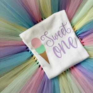 May include: A white t-shirt featuring a graphic of an ice cream cone with a pink scoop and a green scoop, accompanied by the purple text "Sweet one" in a cursive font, layered over a multi-colored pastel tulle skirt with layers of pink, blue, yellow, and green.