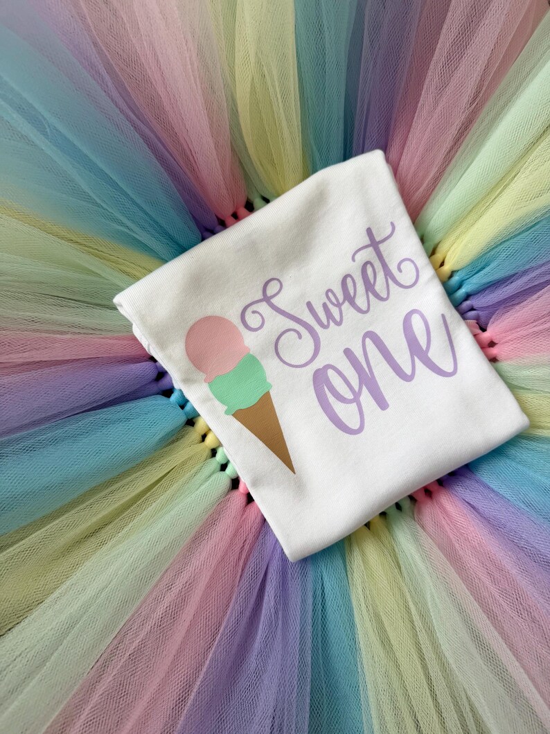 May include: A white t-shirt featuring a graphic of an ice cream cone with a pink scoop and a green scoop, accompanied by the purple text "Sweet one" in a cursive font, layered over a multi-colored pastel tulle skirt with layers of pink, blue, yellow, and green.