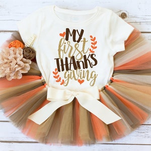 May include: A white onesie with the text "My first THANKSgiving" in gold glitter with red hearts and brown leaves. The onesie is paired with a multi-colored tulle skirt in shades of brown, beige, and orange.