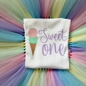 May include: A white t-shirt featuring a graphic of a two-scoop ice cream cone with a pink top scoop and a mint green bottom scoop. The text "Sweet one" is printed in a purple cursive font to the right of the ice cream cone graphic.