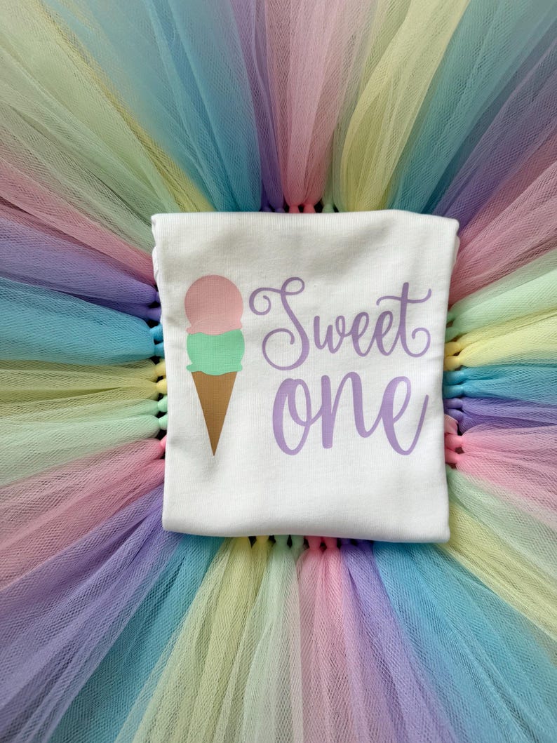 May include: A white t-shirt featuring a graphic of a two-scoop ice cream cone with a pink top scoop and a mint green bottom scoop. The text "Sweet one" is printed in a purple cursive font to the right of the ice cream cone graphic.