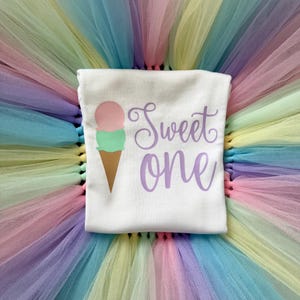 May include: A white t-shirt featuring a graphic of a two-scoop ice cream cone in pink and mint green with a brown cone. The text "Sweet one" is printed in purple script lettering. The shirt is placed on a multi-colored pastel tulle skirt.
