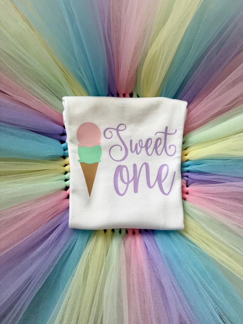 May include: A white t-shirt featuring a graphic of a two-scoop ice cream cone in pink and mint green with a brown cone. The text "Sweet one" is printed in purple script lettering. The shirt is placed on a multi-colored pastel tulle skirt.