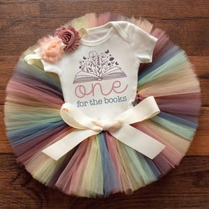May include: A baby outfit featuring a white onesie with a floral and book design, paired with a colorful rainbow tutu. The onesie reads "one for the books." A cream-colored bow and floral headband complete the ensemble.