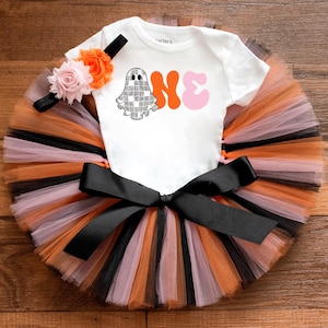 May include: A Halloween baby outfit with a white onesie, a ghost graphic, and the word "ONE". The outfit includes a tutu skirt in orange, black, and pink tulle, a black bow, and a floral headband. The outfit is designed for a baby's first Halloween celebration.