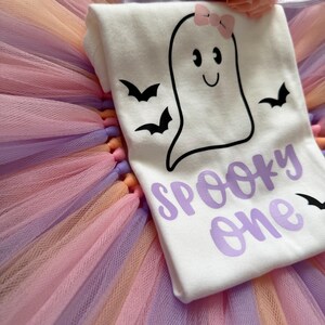 May include: A white bodysuit featuring a black ghost graphic with a pink bow, black bats, and the purple text "SPOOKY one" layered over a multi-coloured tulle tutu skirt in shades of pink, orange, and purple, topped with a glittery crown headband.
