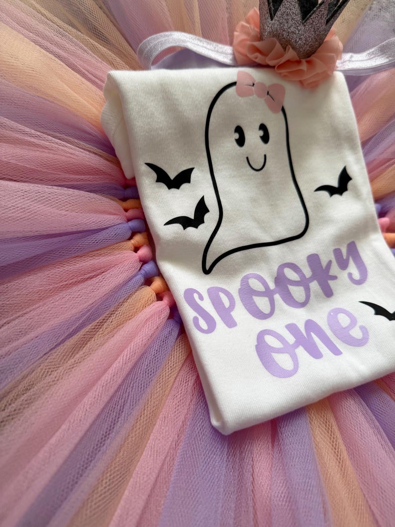 May include: A white bodysuit featuring a black ghost graphic with a pink bow, black bats, and the purple text "SPOOKY one" layered over a multi-coloured tulle tutu skirt in shades of pink, orange, and purple, topped with a glittery crown headband.