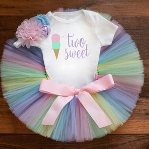 May include: A white onesie with the text "two sweet" and a pink and green ice cream cone graphic. The onesie is paired with a rainbow tulle tutu and a pink bow.