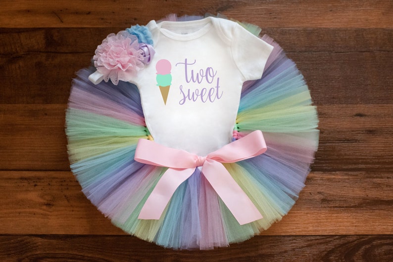 May include: A white onesie with the text "two sweet" and a pink and green ice cream cone graphic. The onesie is paired with a rainbow tulle tutu and a pink bow.