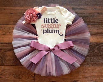 May include: A white baby onesie with the text "little sugar plum" in brown and pink lettering. The onesie is paired with a multi-colored tulle tutu with a pink satin ribbon and a pink and purple flower headband.