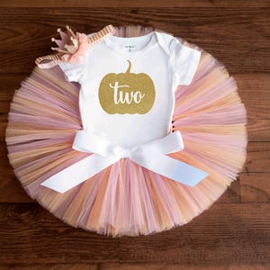 May include: A white short-sleeved bodysuit featuring a gold glitter pumpkin graphic with the word two printed in white script. The bodysuit is paired with a multi-layered tulle tutu in shades of peach and pink, accented by a white satin bow.