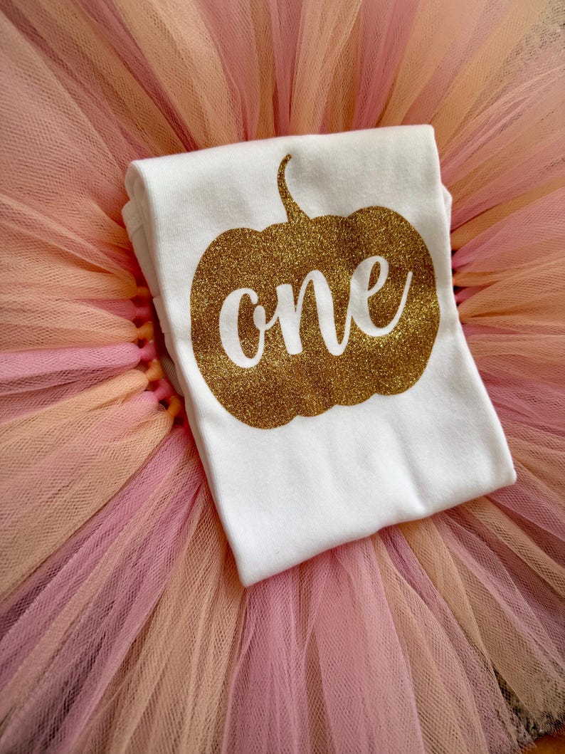 May include: A white t-shirt featuring a gold glitter pumpkin graphic with the word one written in white script across the center, placed on top of a layered tulle skirt in shades of light pink and peach.