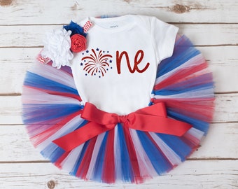 May include: A white baby bodysuit with the word "one" in red script and a firework design. It is paired with a red, white, and blue tutu skirt and a matching headband with floral accents. The outfit is designed for a baby's first birthday.