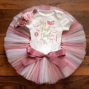 May include: A white short-sleeved bodysuit with the gold and pink text My First Chapter and book-themed graphics, paired with a multi-layered pink and white tulle tutu skirt featuring a large mauve bow and a matching floral headband accessory.