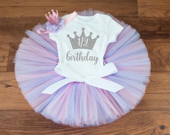 May include: A white onesie with a silver glitter crown and the text "1/2 birthday". The onesie is paired with a pink and purple tulle tutu and a pink crown headband.