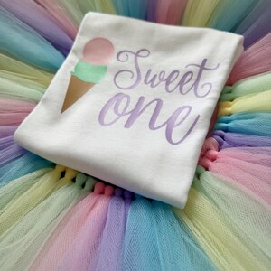 May include: A white t-shirt featuring a graphic of an ice cream cone with a pink scoop and a green scoop, accompanied by the purple script text "Sweet one". The shirt is placed on a multi-colored pastel tulle skirt with layers of pink, blue, and yellow.