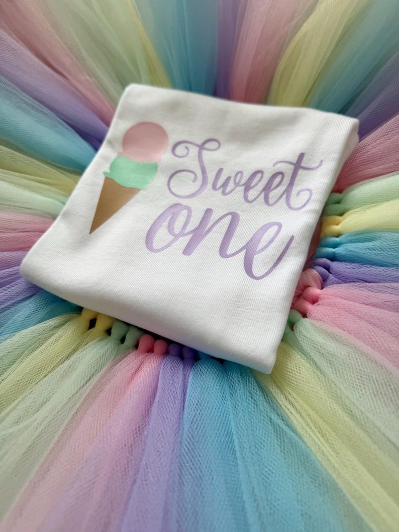 May include: A white t-shirt featuring a graphic of an ice cream cone with a pink scoop and a green scoop, accompanied by the purple script text "Sweet one". The shirt is placed on a multi-colored pastel tulle skirt with layers of pink, blue, and yellow.