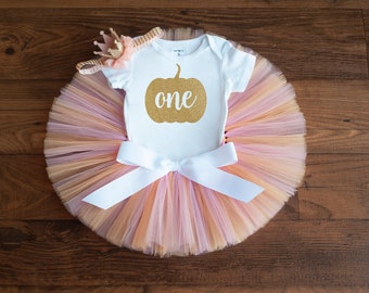 May include: A white onesie with a gold glitter pumpkin and the word "one" on it. The onesie is paired with a peach, pink, and gold tulle tutu and a gold crown headband.