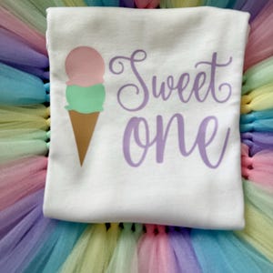 May include: A white t-shirt featuring a graphic of a two-scoop ice cream cone with pink and green scoops and a brown cone. The text "Sweet one" is printed in a purple cursive font to the right of the ice cream graphic on the shirt fabric.