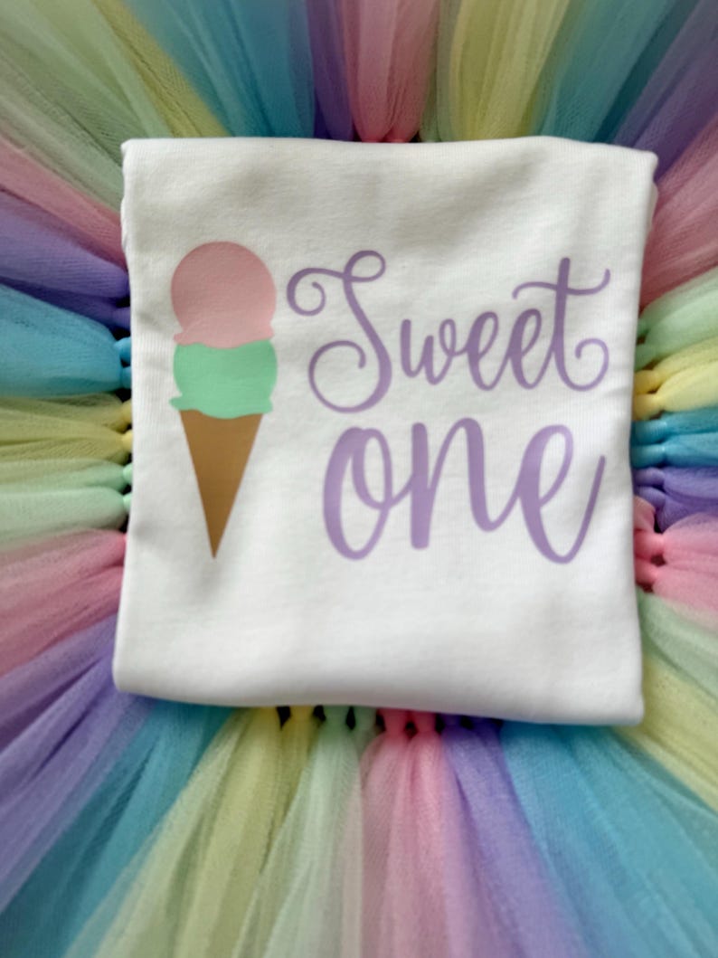 May include: A white t-shirt featuring a graphic of a two-scoop ice cream cone with pink and green scoops and a brown cone. The text "Sweet one" is printed in a purple cursive font to the right of the ice cream graphic on the shirt fabric.