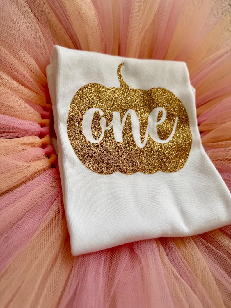 May include: A white short-sleeved t-shirt featuring a gold glitter pumpkin graphic with the word one written in white script across the center. The shirt is placed on top of a layered tulle skirt in shades of light pink, peach, and orange.