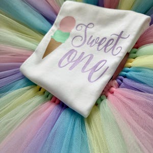 May include: A white t-shirt featuring a graphic of an ice cream cone with a pink scoop and a green scoop, accompanied by the purple script text "Sweet one". The shirt is placed on a multi-colored tulle skirt with layers of pastel pink, blue, and yellow.