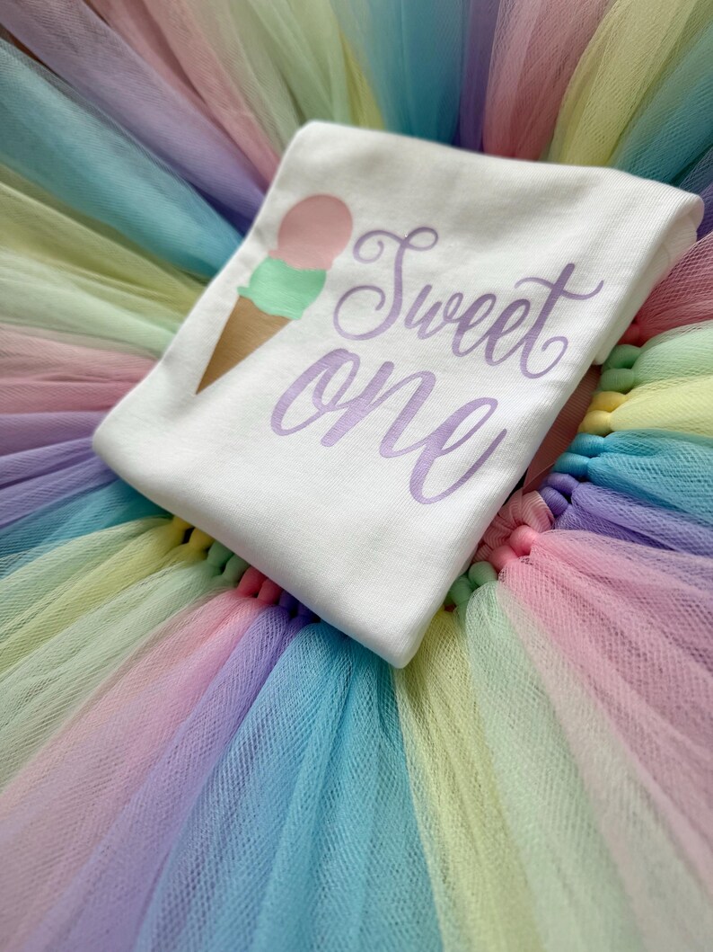 May include: A white t-shirt featuring a graphic of an ice cream cone with a pink scoop and a green scoop, accompanied by the purple script text "Sweet one". The shirt is placed on a multi-colored tulle skirt with layers of pastel pink, blue, and yellow.