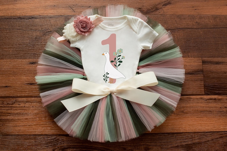 K&ouml;nnte beinhalten: Ein wei&szlig;er Strampler mit einer pinkfarbenen Nummer eins und einer wei&szlig;en Gans mit gr&uuml;nen Bl&auml;ttern. Der Strampler wird mit einem mehrfarbigen T&uuml;ll-Tutu mit einer wei&szlig;en Schleife aus Satin kombiniert.