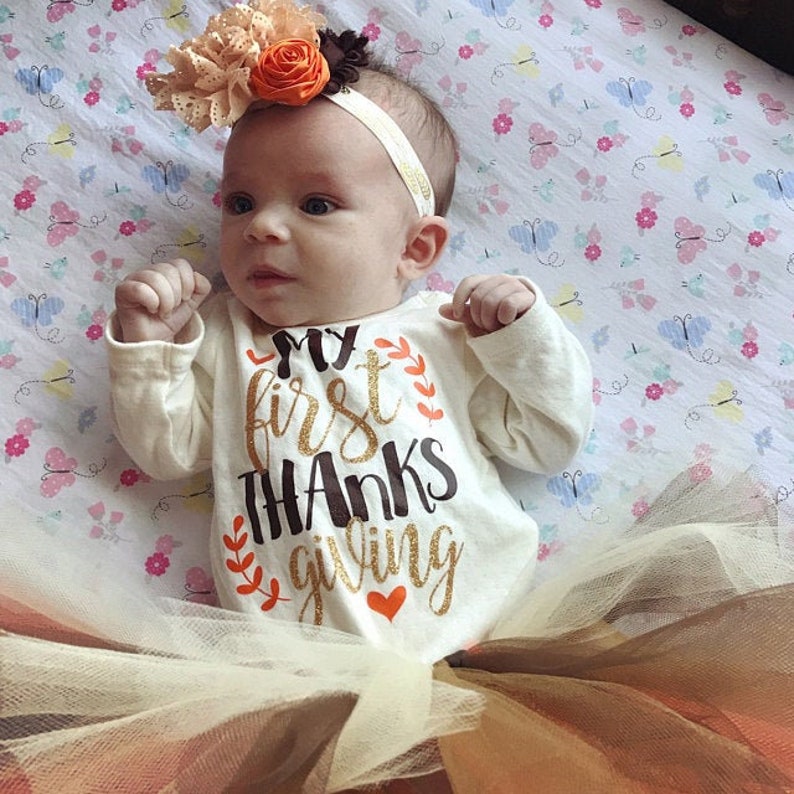 May include: A baby wearing a white long-sleeved onesie with the text "My First Thanks Giving" in gold glitter lettering. The baby is wearing a brown and orange flower headband and a tulle skirt.