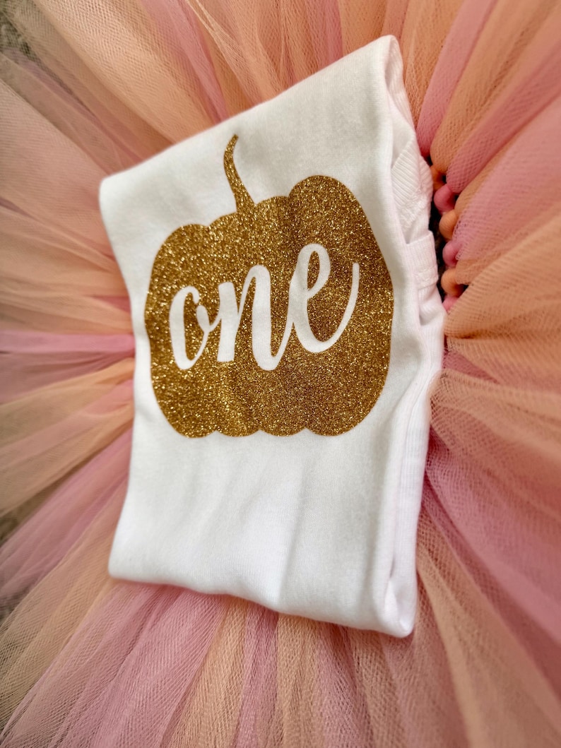 May include: A white short-sleeved shirt featuring a gold glitter pumpkin graphic with the word one written in white script across the center. The shirt is layered over a full, layered tulle skirt in shades of light pink and peach.
