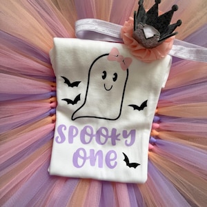 May include: A white short-sleeved shirt featuring a black outline of a ghost with a pink bow, surrounded by black bats and the purple text "SPOOKY one". The shirt is placed on a layered tulle tutu in shades of peach and lavender, with a matching headband.
