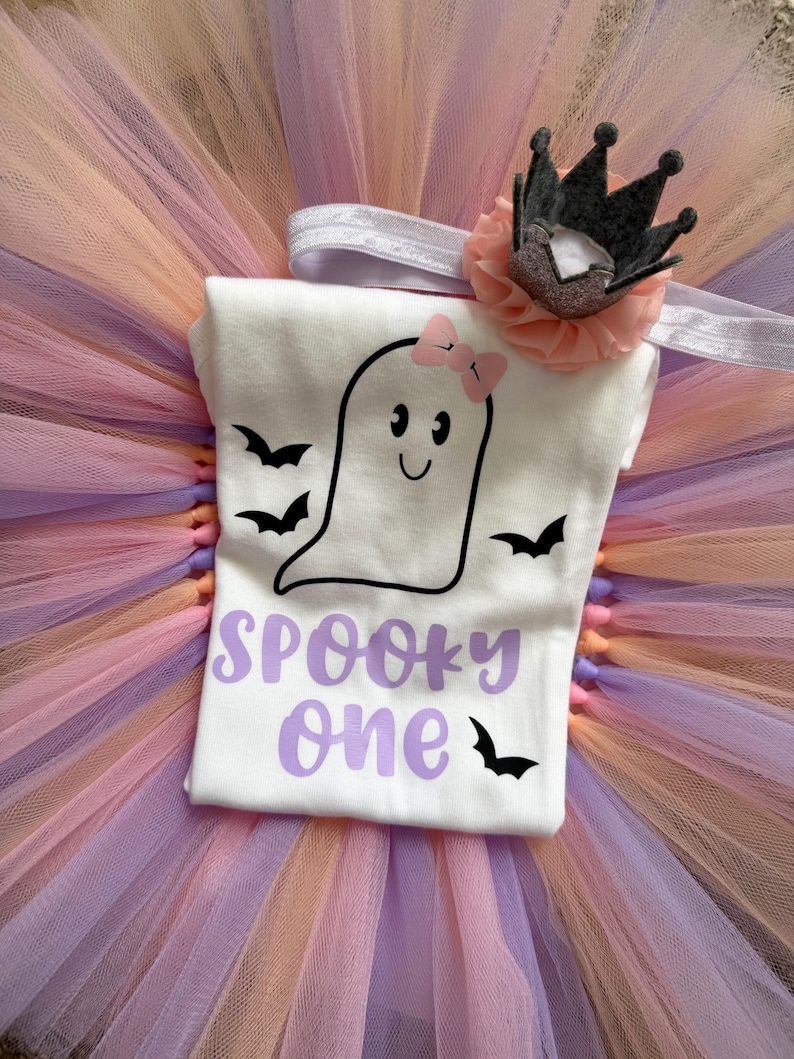 May include: A white short-sleeved shirt featuring a black outline of a ghost with a pink bow, surrounded by black bats and the purple text "SPOOKY one". The shirt is placed on a layered tulle tutu in shades of peach and lavender, with a matching headband.