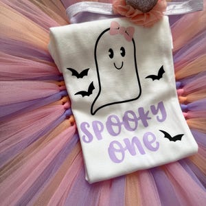 May include: A white t-shirt featuring a black ghost graphic with a pink bow, three black bats, and the purple text "SPOOKY one". The shirt is placed on a multi-coloured tulle tutu skirt in shades of orange, purple, and pink, accompanied by a headband.