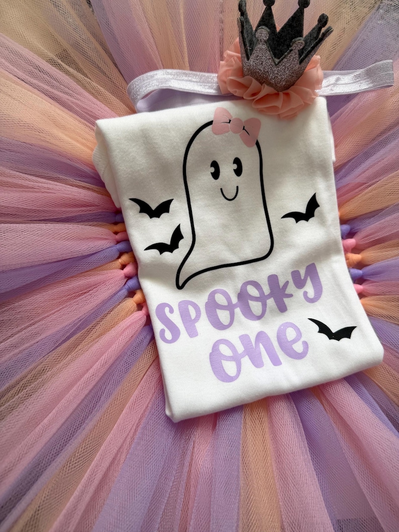 May include: A white t-shirt featuring a black ghost graphic with a pink bow, three black bats, and the purple text "SPOOKY one". The shirt is placed on a multi-coloured tulle tutu skirt in shades of orange, purple, and pink, accompanied by a headband.