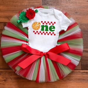 May include: A white baby onesie with a pizza graphic and the word "one" in green, set on a red, white, and green tutu. A red bow and a floral headband complete the outfit. The outfit is designed for a baby's first birthday.
