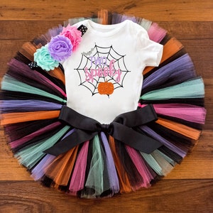 May include: A white onesie with a spiderweb and the text "Too Spooky" in pink and black. The onesie is paired with a rainbow tulle tutu and a flower headband.